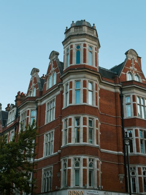 The image features a historic red brick building with multiple bay windows and decorative white stone accents. The structure has a prominent corner tower topped with a rounded turret and a decorative 