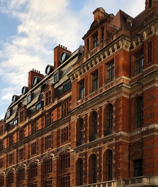 The image shows the upper floors of a historic red-brick building with ornate architectural details, including arched windows, decorative cornices, and dormer windows on the roof. The exterior surface appears clean and well-maintained, with a bright sky in the background. The sunlight highlights the texture of the brickwork and the details of the building's design. Visible tools such as a cloth and spray bottle are placed on a window ledge, indicating ongoing surface cleaning or maintenance activities. The setting suggests a residential or commercial property in an urban area, where professional cleaning and surface sanitisation might be performed by services like Cleaners Knightsbridge to maintain hygiene and aesthetic appeal.