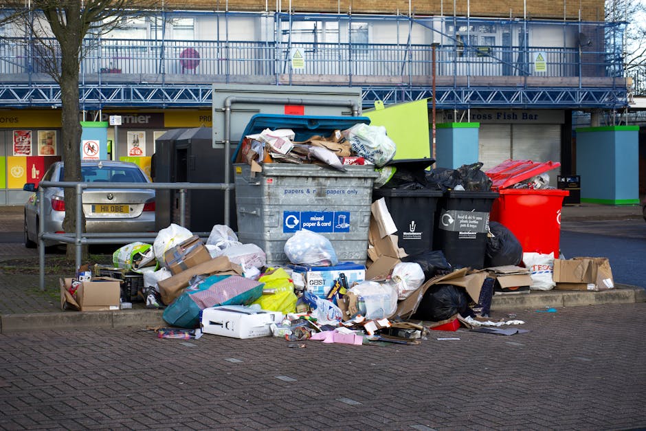 A construction vehicle is parked on a dirt road beside a large pile of bulky waste, including plastic bags, cardboard, and dried plant matter. The surface of the road appears uneven and dirty, with sc